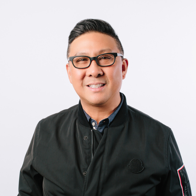A man with short hair and glasses smiles confidently, wearing a black jacket over a collared shirt, against a white background.