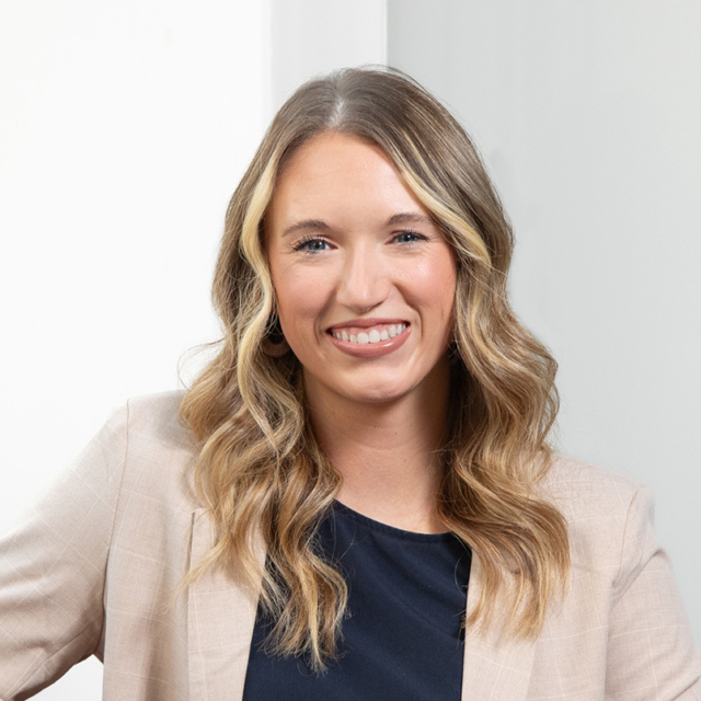 Smiling woman with wavy shoulder-length blonde hair and highlights, wearing a navy top and beige blazer against a pale neutral background.