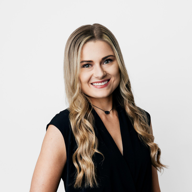 Smiling young blonde woman with long wavy hair wearing a sleeveless black top and small black beaded choker, posed against white.