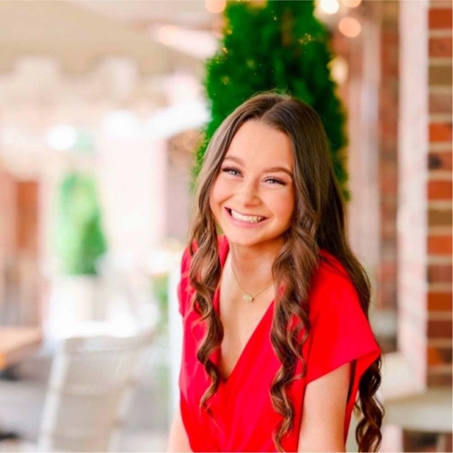 A young woman with long, wavy hair smiles brightly while wearing a red dress, surrounded by soft lighting and greenery in a bright setting.