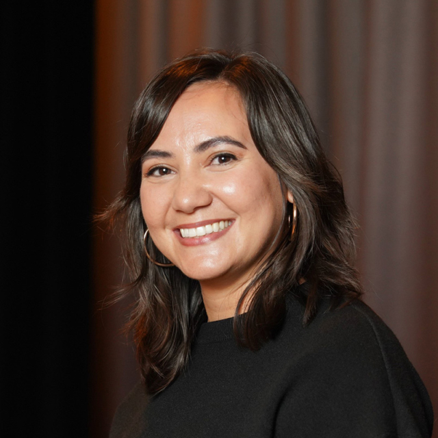 Smiling woman with shoulder-length dark hair, gold hoop earrings, wearing a black top against a soft brown and black curtain backdrop.