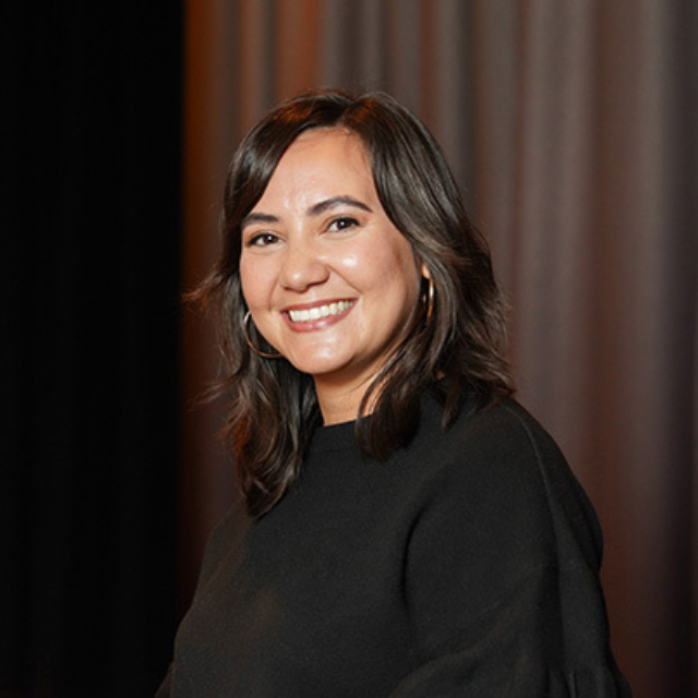 A smiling woman with dark, wavy hair and hoop earrings poses against a softly lit backdrop, wearing a black sweater.