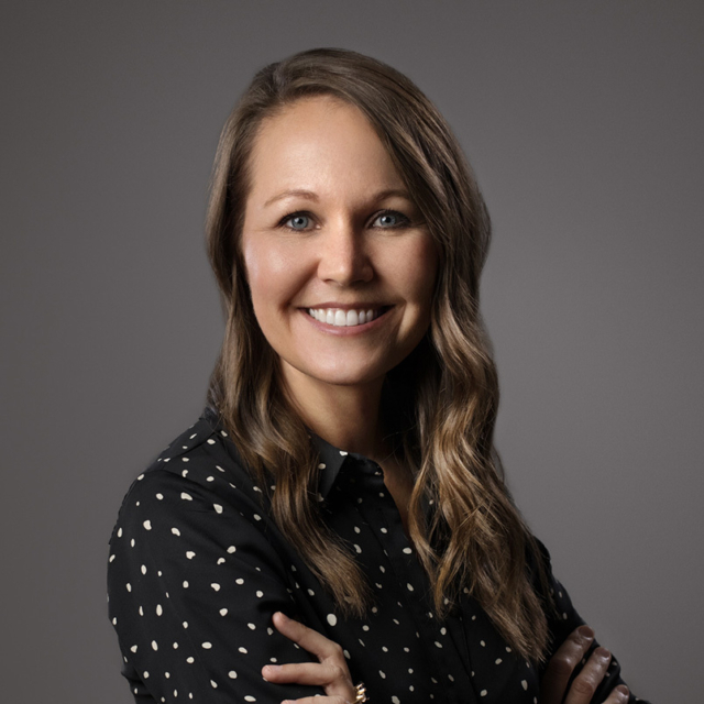 Smiling professional woman with wavy light-brown hair and blue eyes, wearing a black polka-dot blouse with arms crossed against gray.
