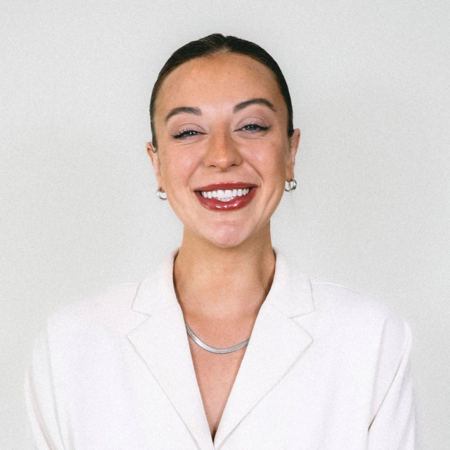 A smiling woman with medium-length hair, wearing a light blazer and hoop earrings, poses against a soft gray background.