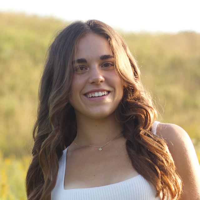 Young woman with long wavy brown hair and a warm smile wearing a white ribbed tank and small gold pendant, sunlit grassy field behind her.