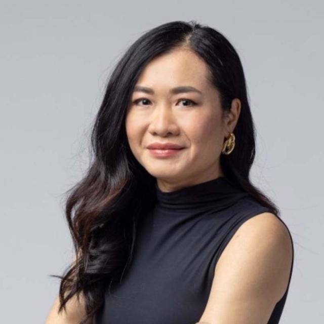 A woman with long, dark hair wears a black sleeveless top and gold earrings, smiling confidently against a neutral background.