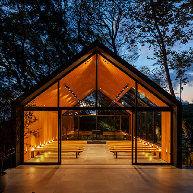 A glass A-frame chapel with warm wooden interior, rows of simple benches and soft amber lighting, set among tall trees at dusk.