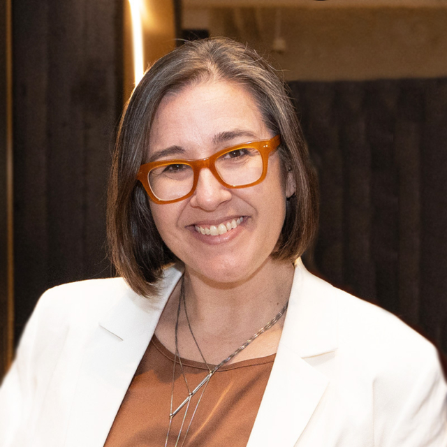 A smiling woman with short hair wears orange glasses and a white blazer over a brown top, standing against a dark background.