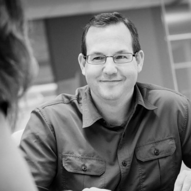 A smiling man with glasses is engaged in conversation, sitting at a table, wearing a collared shirt in a softly lit setting.