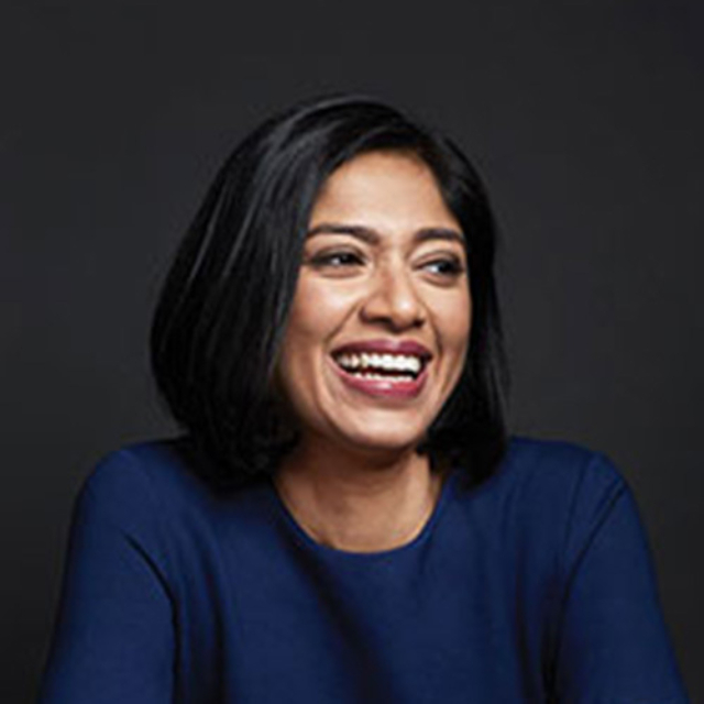 A woman with shoulder-length black hair smiles brightly while wearing a blue top, set against a dark background.