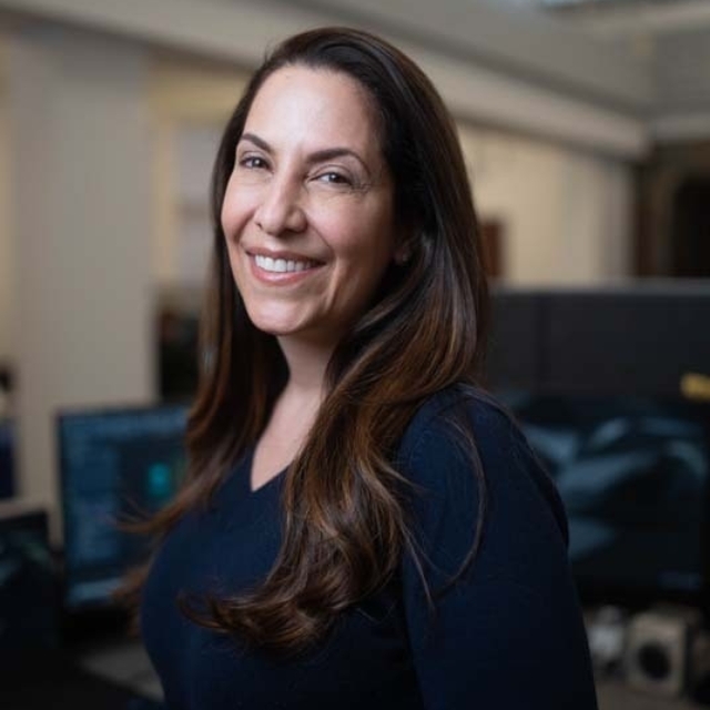 A woman with long, dark hair smiles warmly while standing in an office with computer monitors visible in the background.