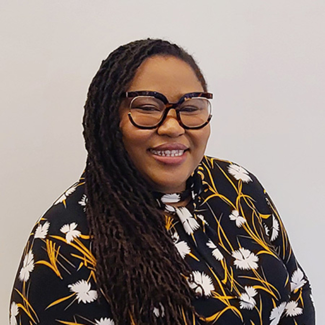 A woman with long, dark, dreadlocked hair smiles warmly while wearing large glasses and a floral black and yellow blouse against a plain background.