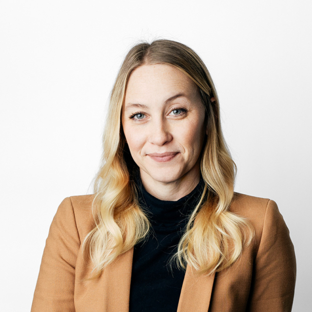 Head-and-shoulders portrait of a blonde woman with wavy hair, blue eyes and a subtle smile, wearing a camel blazer and black turtleneck.