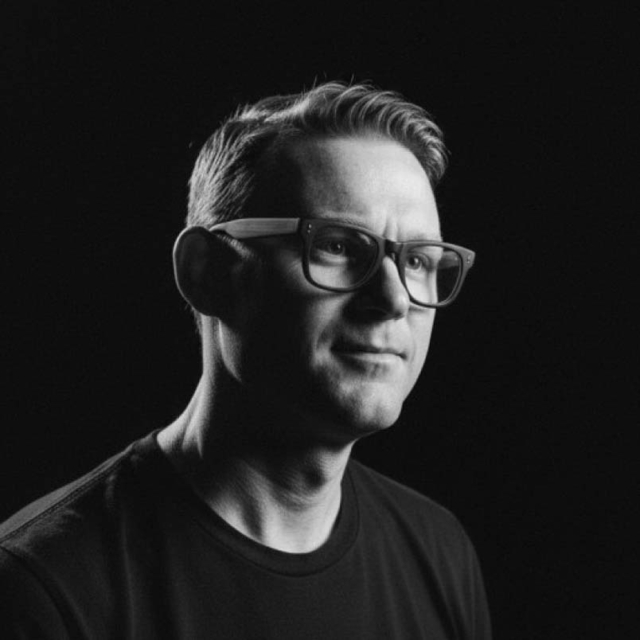 A man with light hair and glasses gazes thoughtfully to the side, set against a dark background that highlights his features.