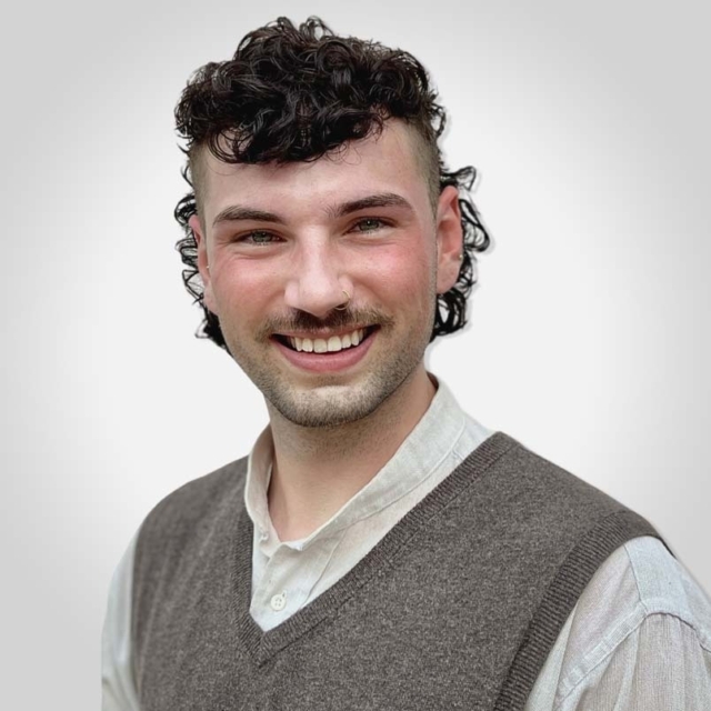 A person with curly hair and a mustache smiles while wearing a light-colored shirt and a gray vest against a soft gradient backdrop.