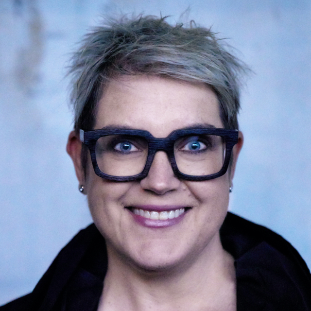 A woman with short, spiked hair and oversized black glasses smiles warmly, wearing a black top against a soft blue backdrop.
