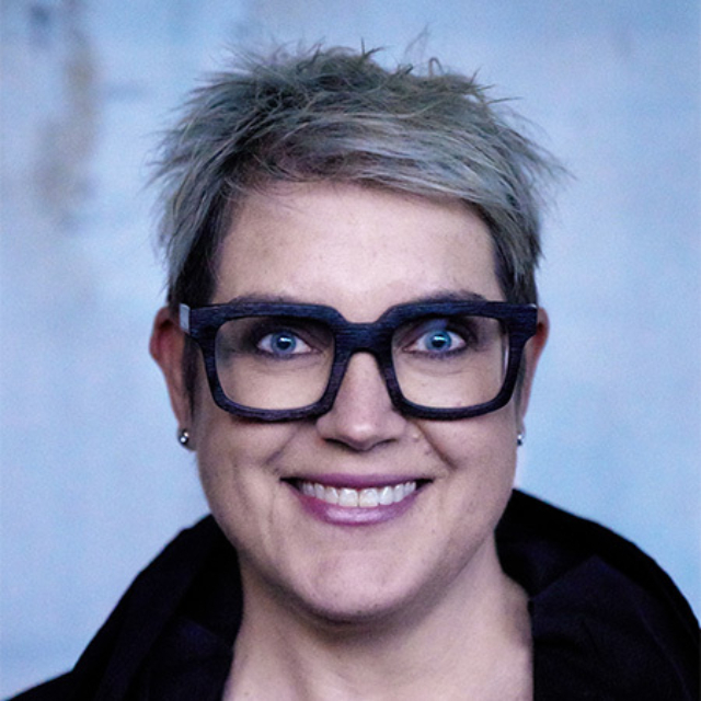 A person with short, spiky hair and large black glasses smiles warmly against a soft, blurred background, emanating a friendly vibe.