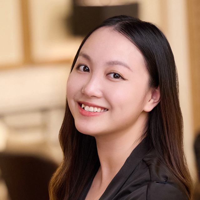 Smiling young woman with long dark hair and winged eyeliner, wearing a black blouse, head turned to camera against warm blurred interior.