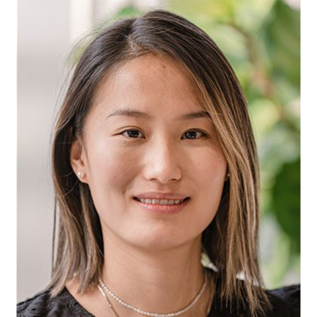 A young woman with medium-length hair smiles softly, wearing a black top and delicate jewelry, against a blurred green backdrop.
