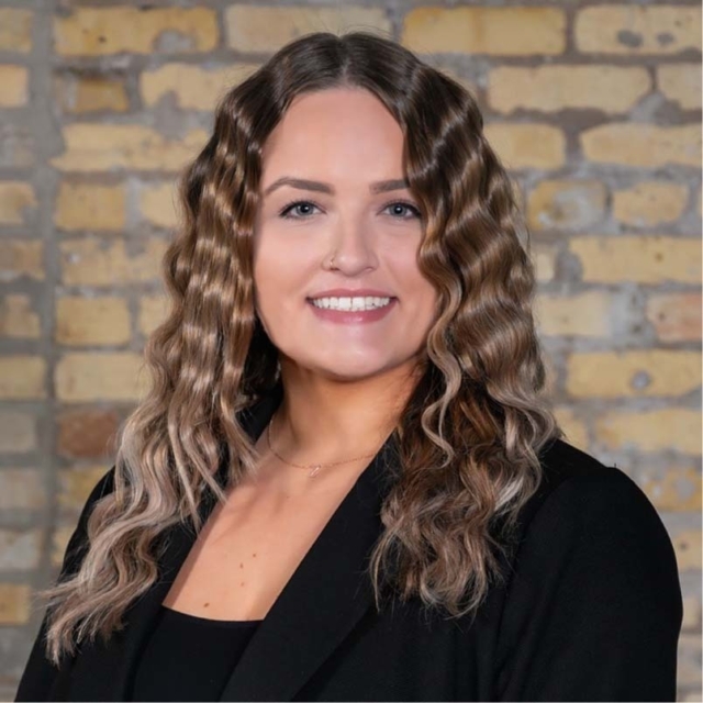 A woman with wavy, shoulder-length hair smiles warmly, wearing a black blazer against a textured yellow brick background.