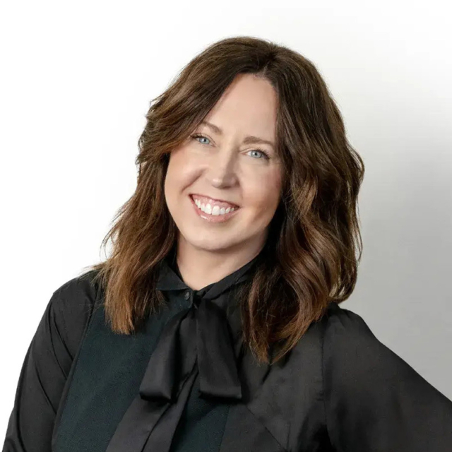 Smiling woman with shoulder-length wavy brown hair, blue eyes, and white teeth, wearing a black blouse with a bow collar against pale gray.