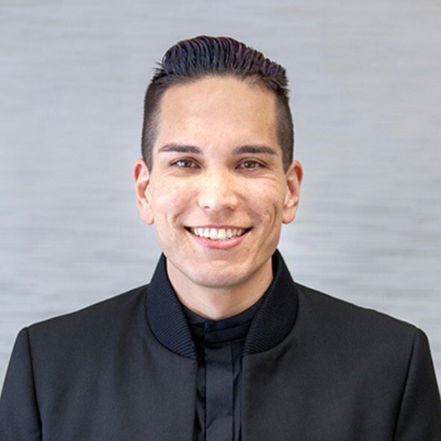 A smiling individual with short, slicked-back hair wearing a dark jacket. The background features a subtle, textured gray pattern.