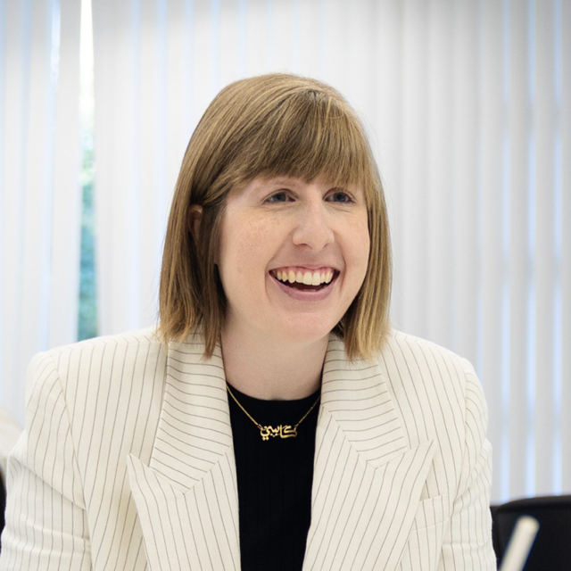 Smiling woman with short light brown hair in a white pinstripe blazer, black top and gold pendant with Arabic script, blinds behind her.