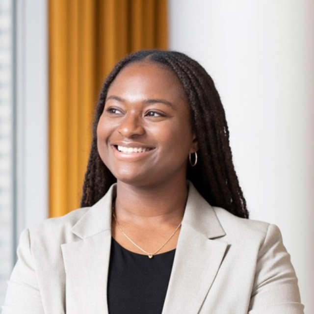 Smiling woman with long braided hair, light gray blazer over black top, gold pendant necklace and hoop earring, warmly gazing left.