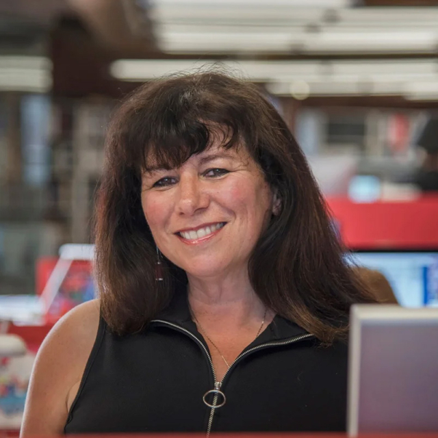 Smiling woman with shoulder-length dark hair and bangs wears a sleeveless black zippered top with a round zipper pull, seated in red office.
