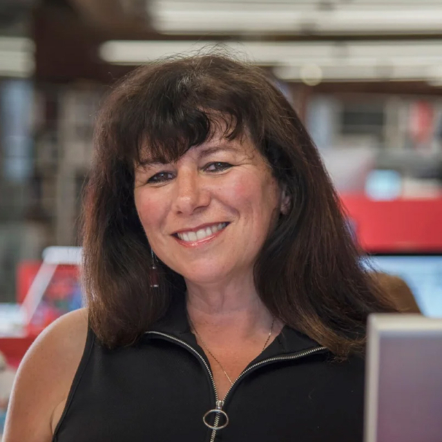 Smiling middle-aged woman with dark shoulder-length hair and bangs, wearing a black zip-top with circular zipper pull, library background.