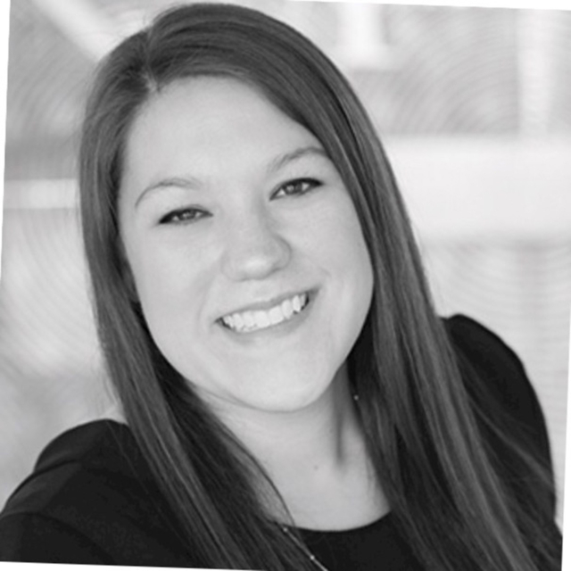 A woman with long brown hair smiles warmly, wearing a black top, in front of a soft-focus background with light patterns.