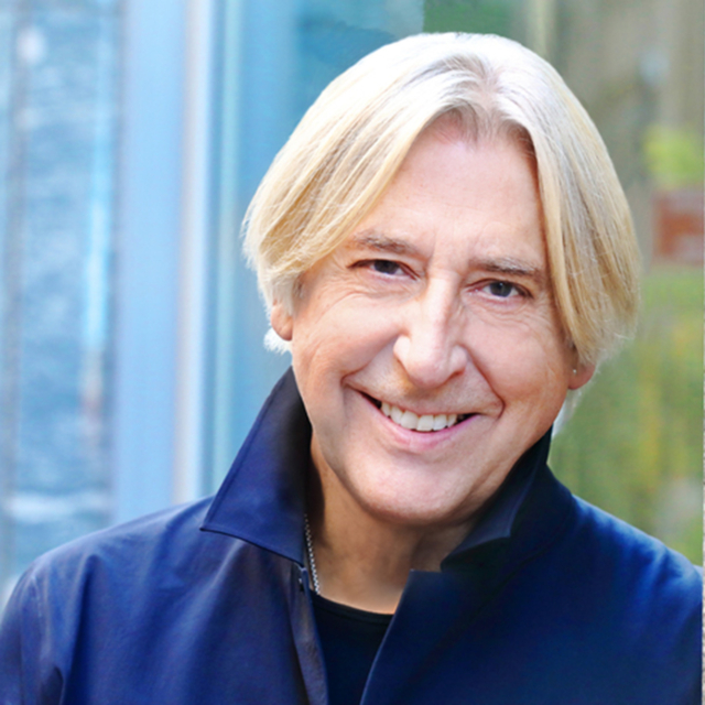 A smiling individual with shoulder-length blonde hair wears a dark jacket, standing against a softly blurred outdoor background.