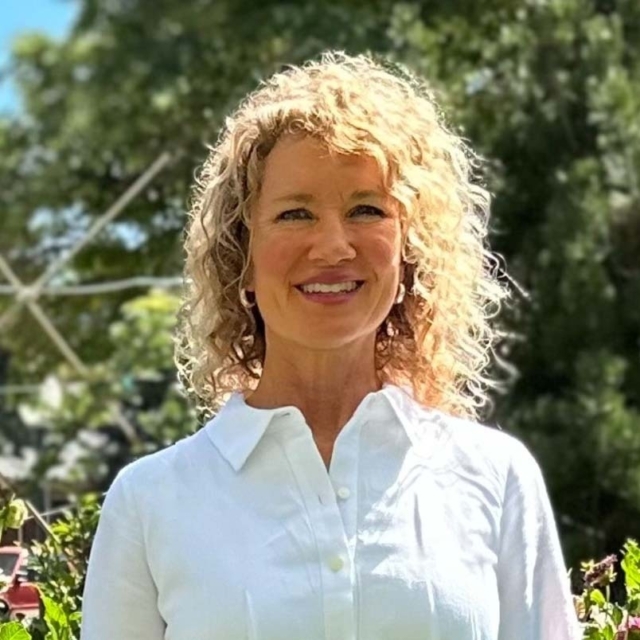 A woman with curly blonde hair smiles warmly, wearing a white shirt, surrounded by greenery and soft, natural light.