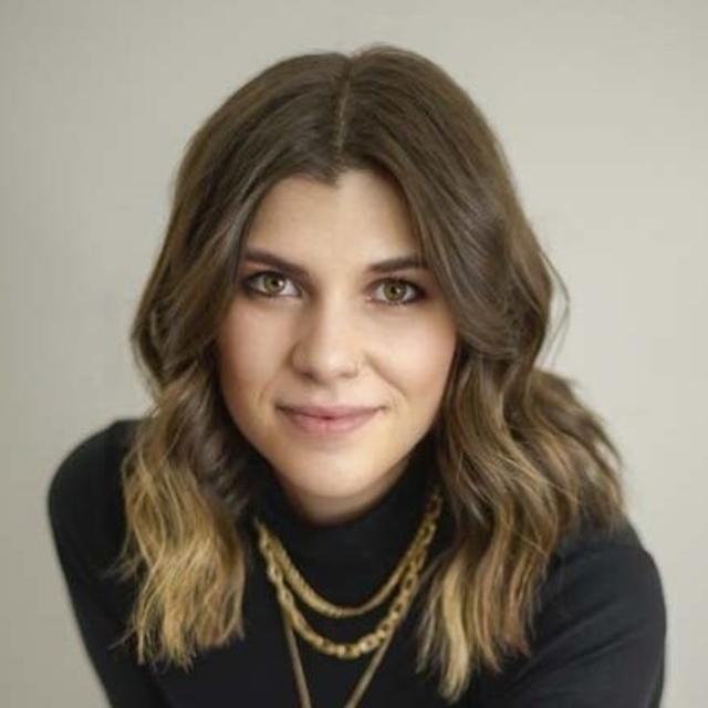 A woman with wavy brown hair poses confidently while wearing a black turtleneck and layered gold necklaces against a light backdrop.