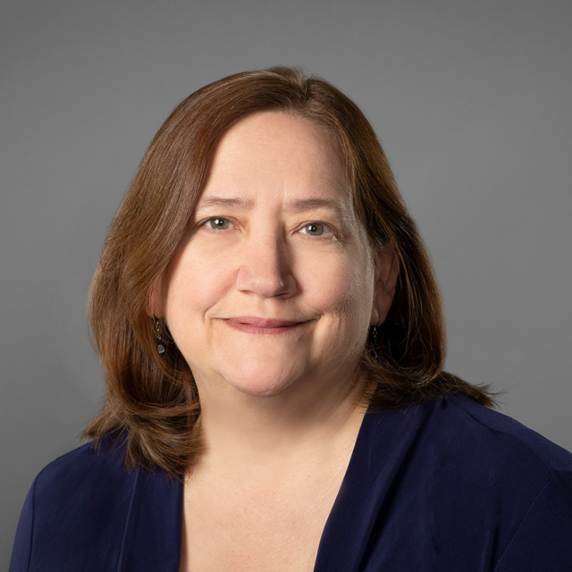 A middle-aged woman with light brown shoulder-length hair in a navy blouse and small earrings, smiling softly against a gray background.