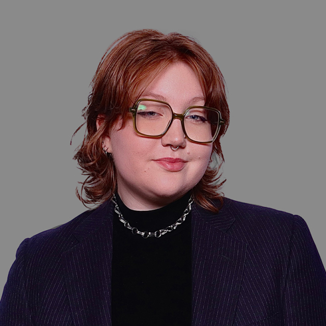 Person with short wavy reddish-brown hair, green oversized glasses, septum ring, subtle smile, chain necklace and pinstriped blazer on gray.