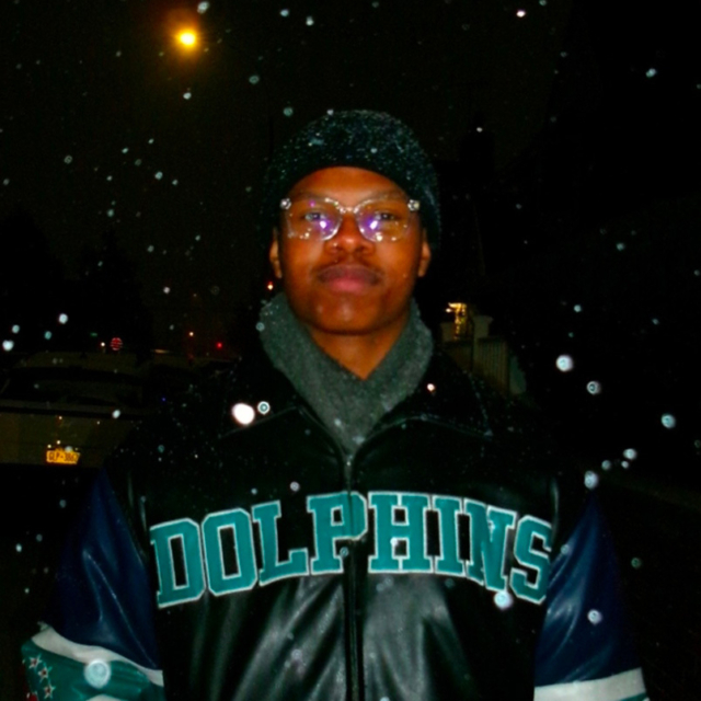 Young person standing outdoors at night in falling snow, wearing a black hat, glasses, gray scarf and a leather jacket reading 'DOLPHINS'.