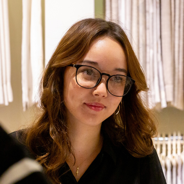Young woman with wavy brown hair wearing black round glasses, small hoop earring, nose ring and cross necklace, smiling in a clothing store.