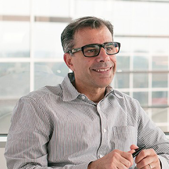 A smiling man with glasses wearing a striped shirt sits indoors, holding a pen, with a bright, modern workspace in the background.
