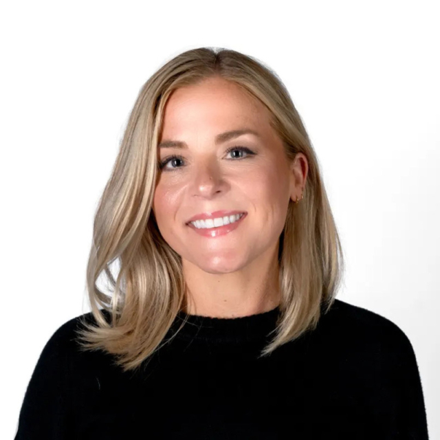 Smiling blonde woman with shoulder-length hair, light makeup and a small hoop earring, wearing a black sweater against a white backdrop.