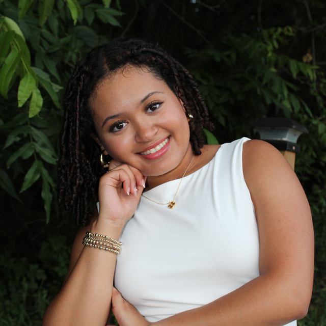 Young woman with short twists smiles, leaning her head on her hand, wearing a white top and gold jewelry against leafy green foliage.
