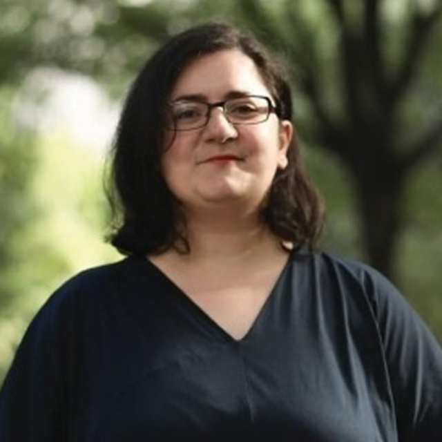 A woman with shoulder-length dark hair and glasses stands confidently outdoors, wearing a navy top, with greenery in the background.