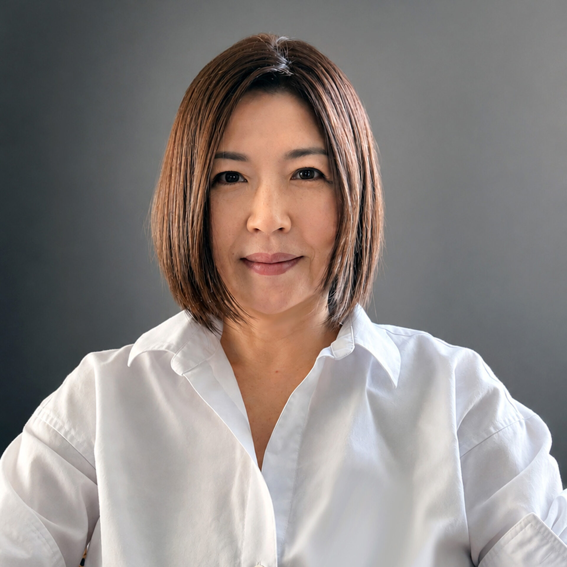 Middle-aged Asian woman with straight chin-length brown bob haircut, wearing a white collared shirt against a neutral gray background.