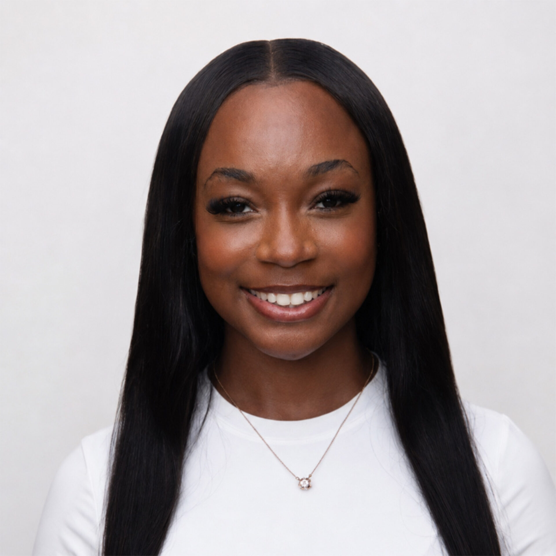 Smiling young woman with long center-parted straight hair, long lashes, white top and small pendant necklace on a pale backdrop.