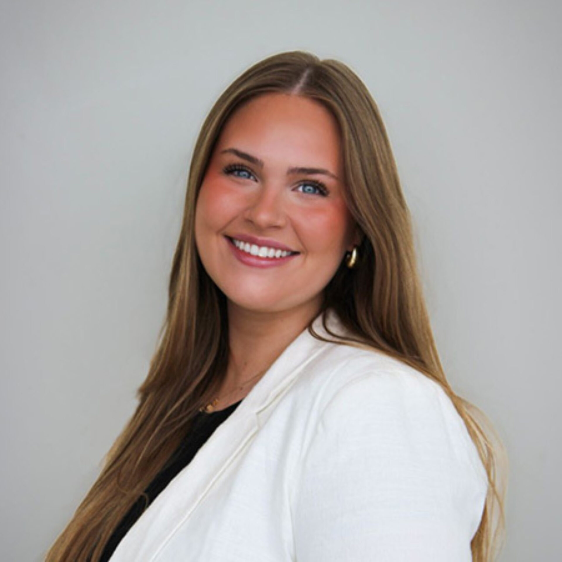 A young woman with long, straight brown hair and blue eyes smiles warmly, wearing a white blazer over a black top against a light background.