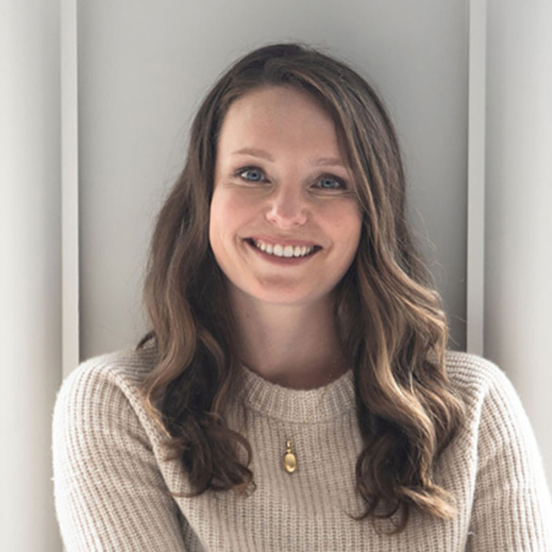 A smiling woman with long, wavy hair, wearing a light sweater, poses in front of a neutral background, radiating warmth and friendliness.