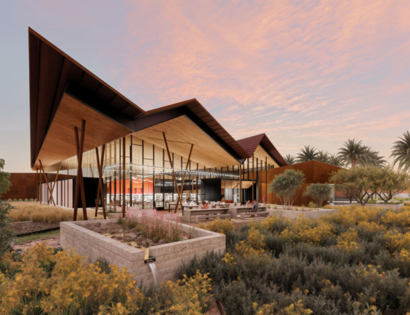 A modern architectural structure with a distinctive angular roof sits amidst lush landscaping, featuring flowering plants and palm trees.