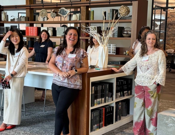 A group of five women engages in cheerful conversation, standing in a stylish workspace surrounded by decorative items and books.