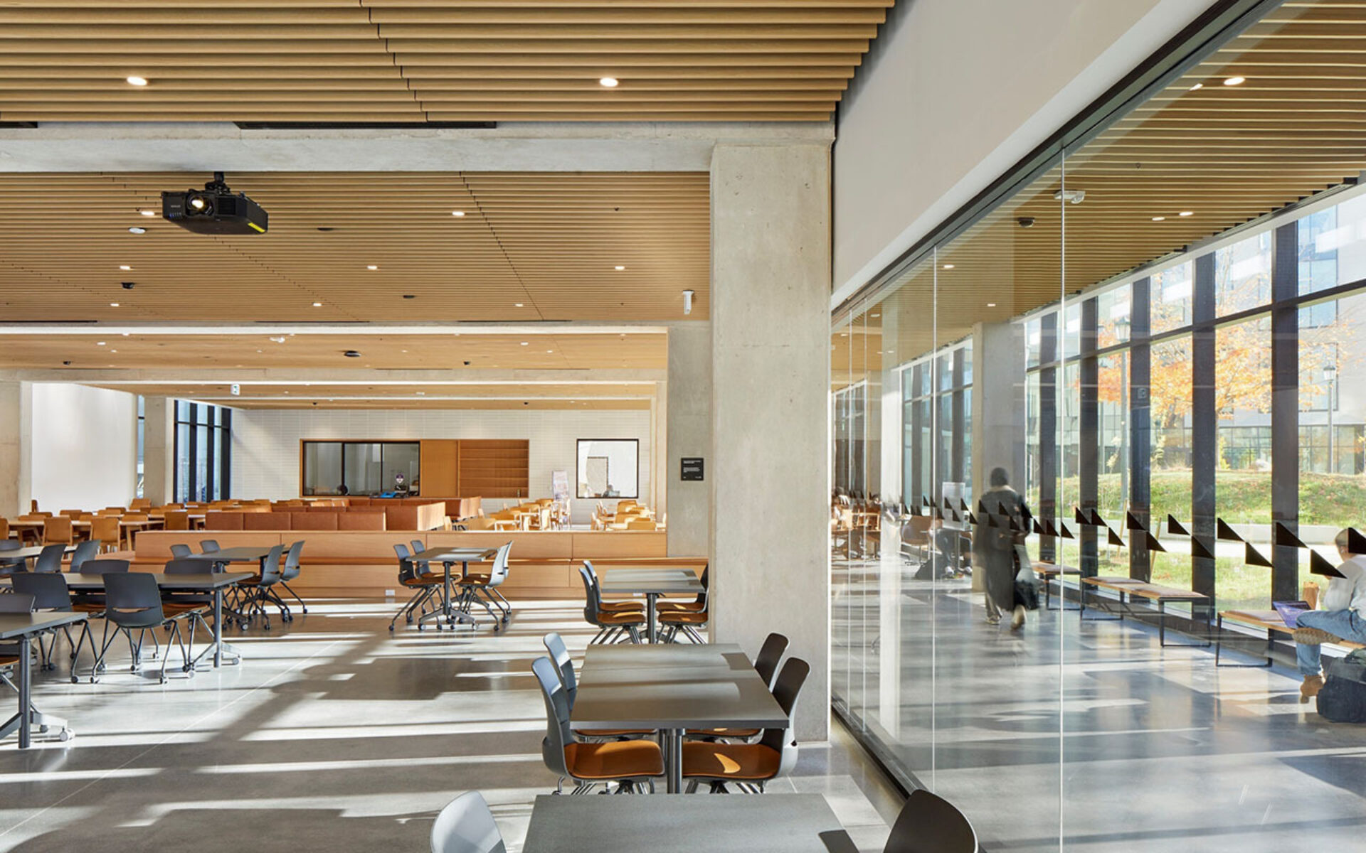 Spacious modern study hall with wooden slatted ceiling, concrete columns, tables and chairs, and a sunlit glass corridor with people.