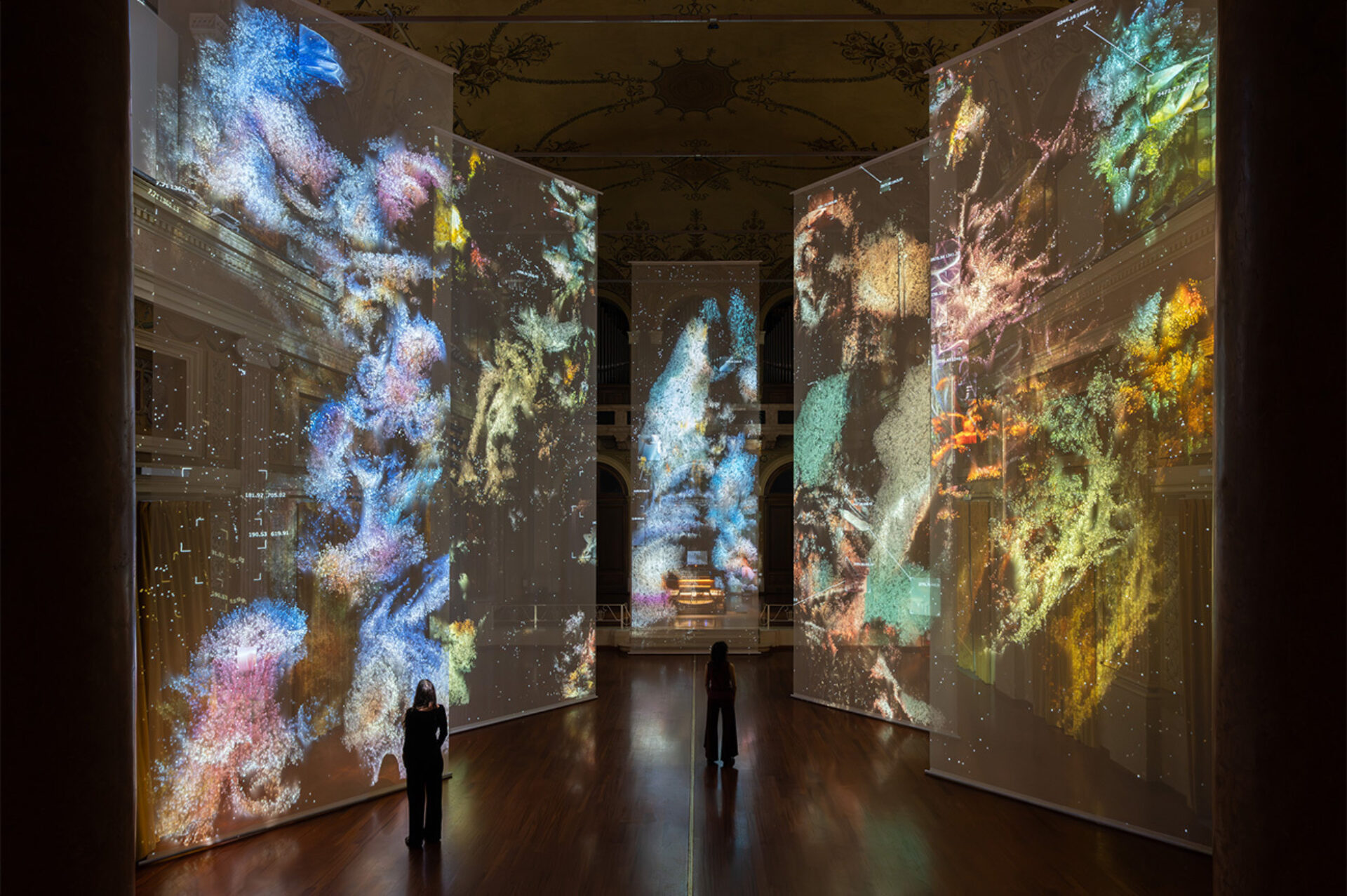 A grand ornate hall with four tall sheer screens showing colorful cosmic projections, two silhouetted figures stand on polished wood floor.